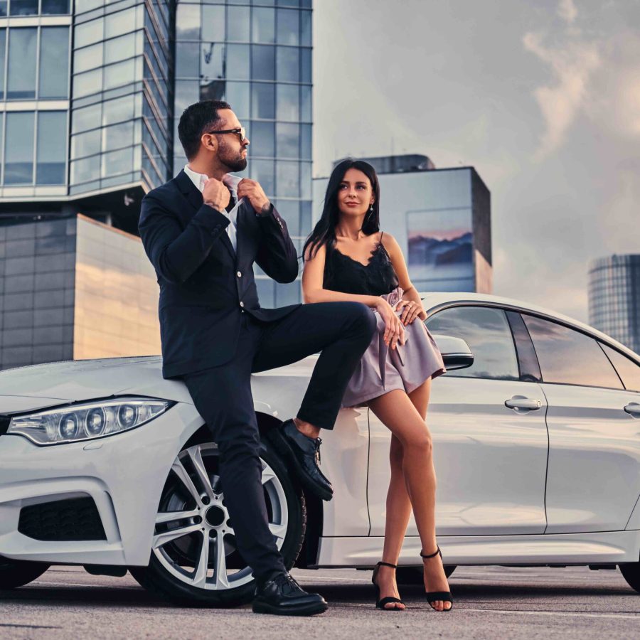 Well-dressed attractive couple leaning on a luxury car outdoors against a skyscraper.
